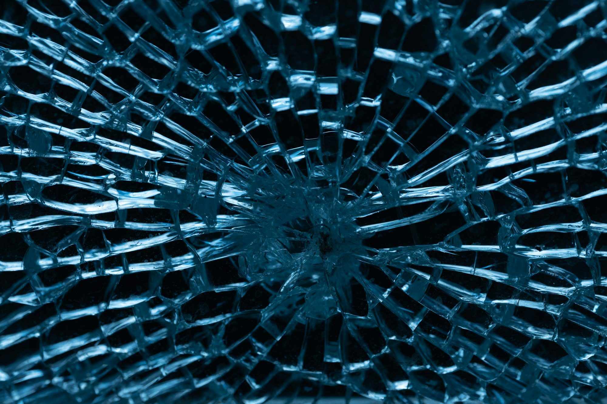 Close-up of a shattered glass pane with cracks radiating out from an impact point, lit blue-green against a dark background