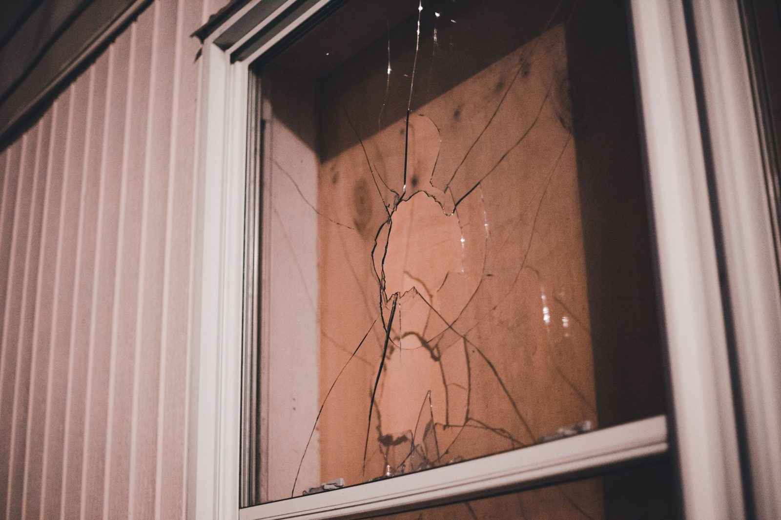 A window pane in a white frame with cracks spreading from an impact point, warm light showing through from behind