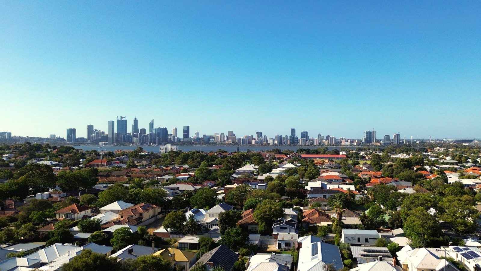 Aerial view across leafy Perth suburban rooftops towards the city skyline under a clear blue sky