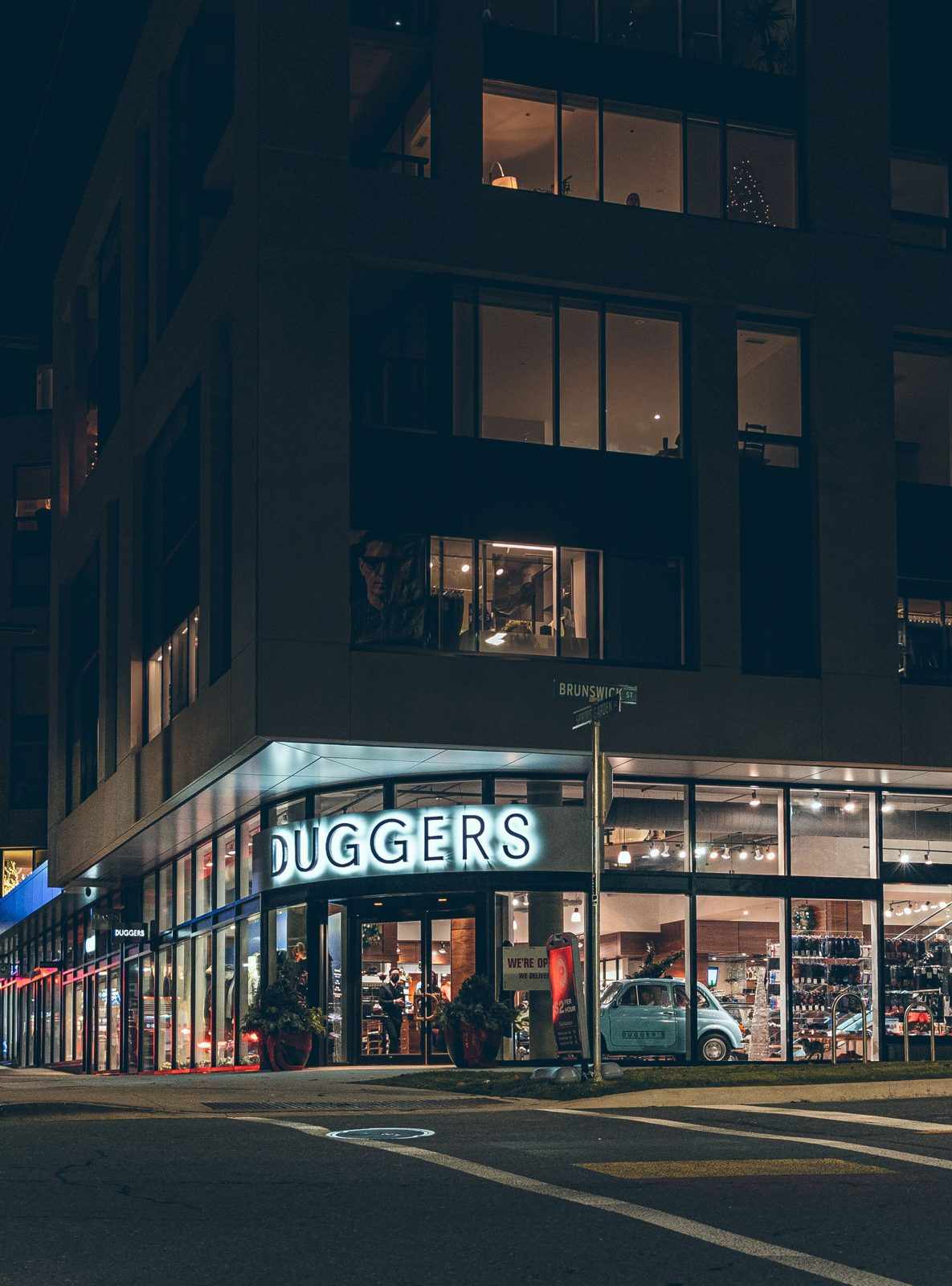 A brightly lit glass shopfront on a city street corner at night, under a dark multi-storey building
