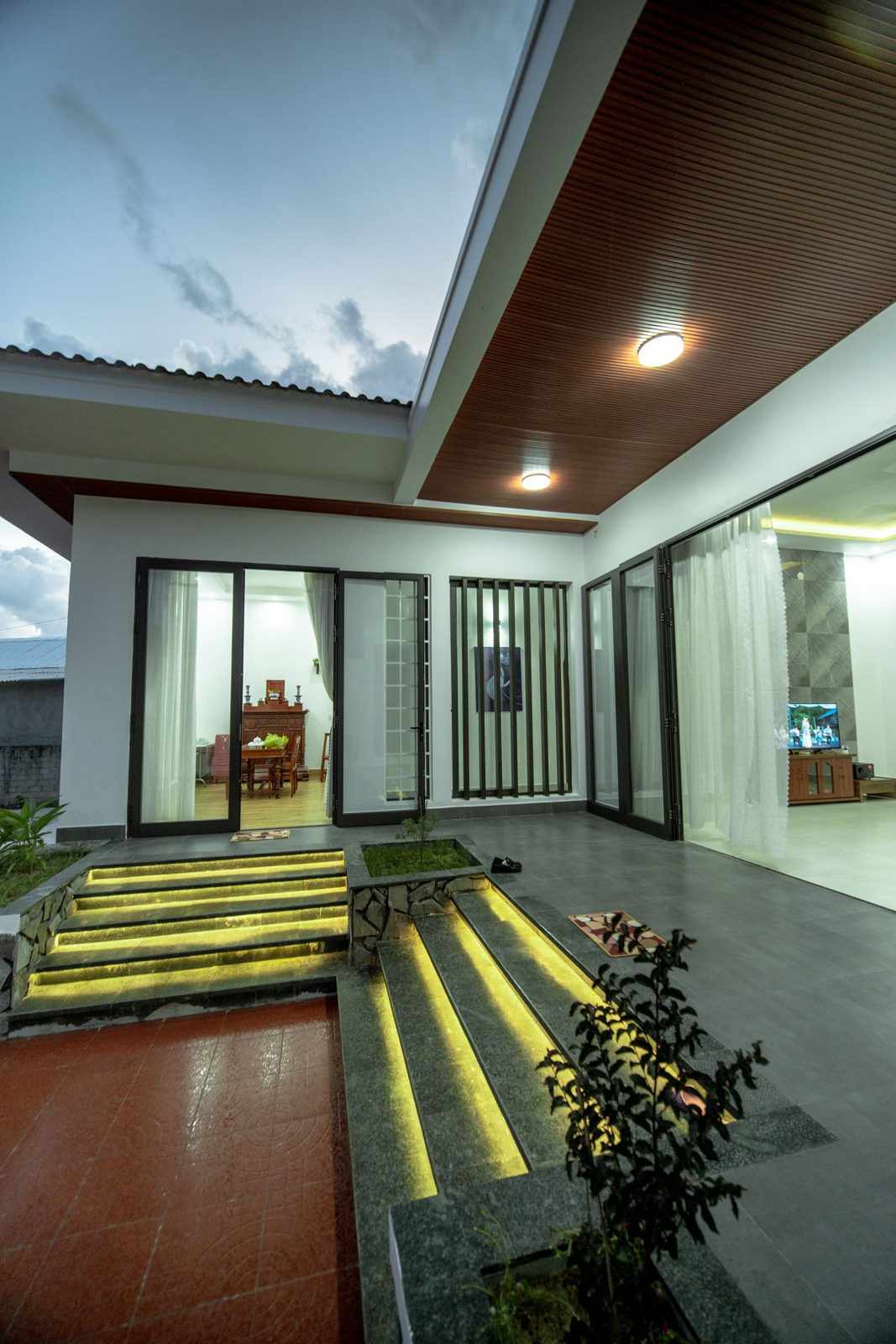 A row of tall glass doors along a covered outdoor area at night, with lit steps and plants in front